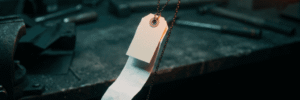 A blank price tag hanging on a chain above a workbench with a long paper receipt unspooling into shadow, illustrating the real cost of AI software for small business