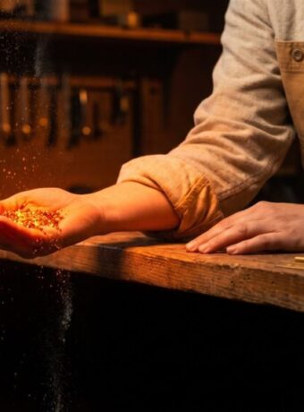 A shop owner's cupped hand catches a heap of glowing orange embers above a workbench while a few slip past her fingers and fall over the counter edge into the dark.