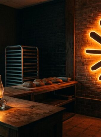 A bakery before dawn with a Claude starburst sign on the brick wall, half its glow gone dark, while an oil lantern burns on the counter as a backup light